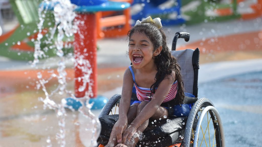 Little girl in wheel chair laughs as she sees the water shoot up in front of her at Morgan’s Wonderland